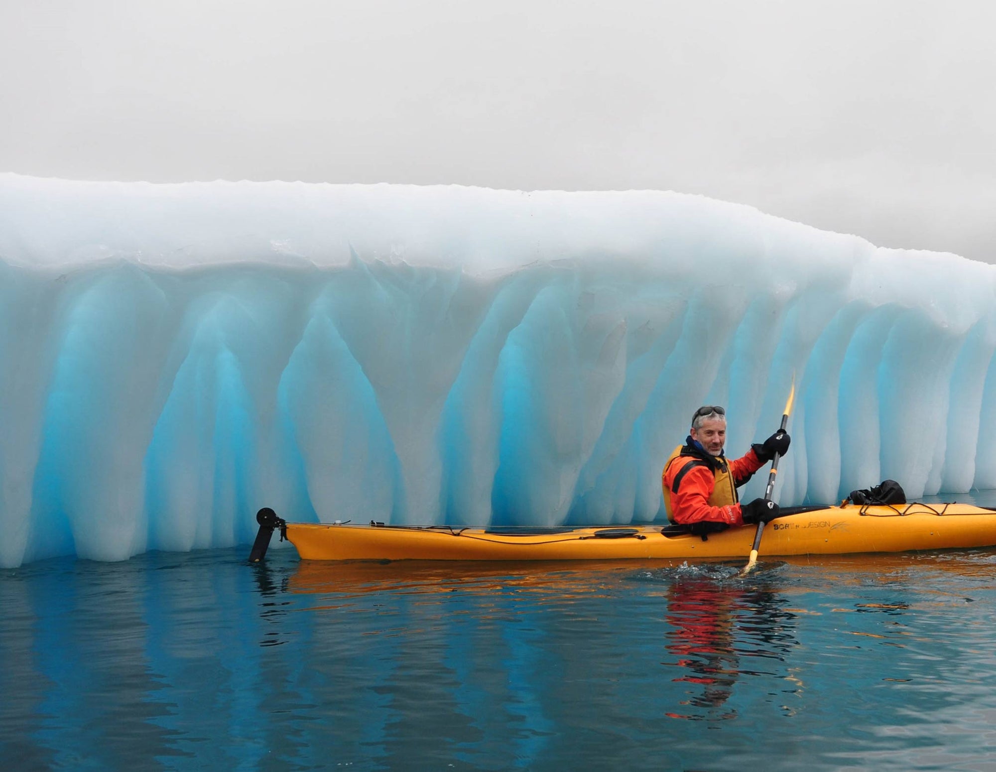 Artist Nicholas Jones in an orange drysuit, kayaking in front of a luminous fluted iceberg off Baffin Island, Canada.