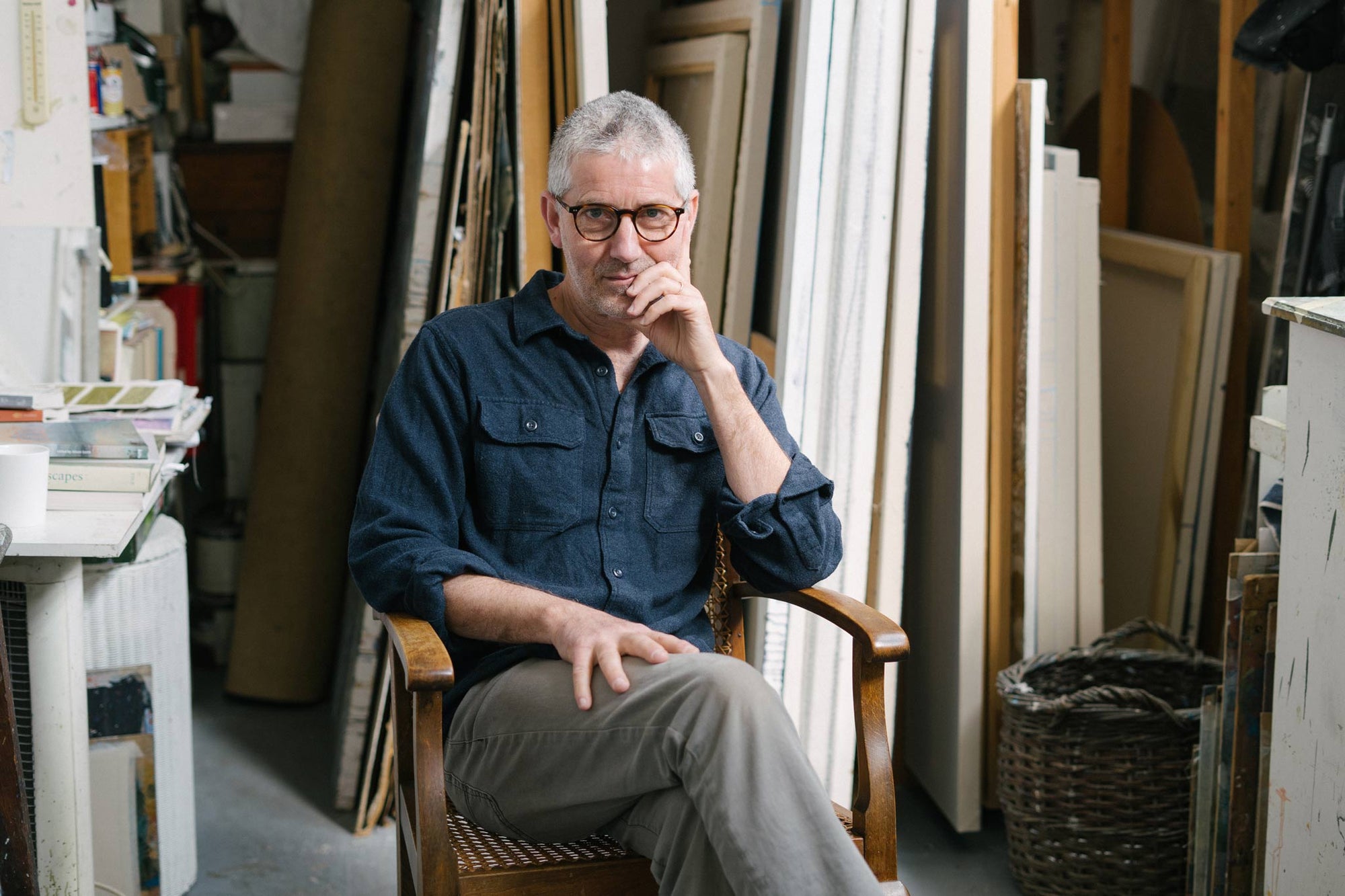 Portrait of artist Nicholas Jones in his studio, seated in an antique chair in front of a storage rack filled with large canvases.
