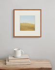 The print “A Hot Wind” hangs above a wooden bench with a book and cup, suggesting the shimmer and tension of heat moving across the land.