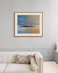 Framed art print of "A Deeper Meeting, Autumn Rainbow Over Holy Isle" by Nicholas Jones, shown in an elegant, minimalist room with a classic sofa and a window seat. Displayed here in size extra-large.