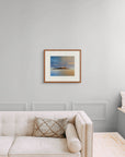 Framed art print of "A Deeper Meeting, Autumn Rainbow Over Holy Isle" by Nicholas Jones, shown in an elegant, minimalist room with a classic sofa and a window seat. Displayed here in size medium.