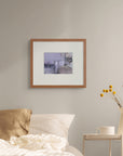 Framed art print of "Cloud, Sea, Snow, Rain", displayed above a bed with light filtering through a gauze curtain. A white jug of yellow flowers stands on a small table nearby.