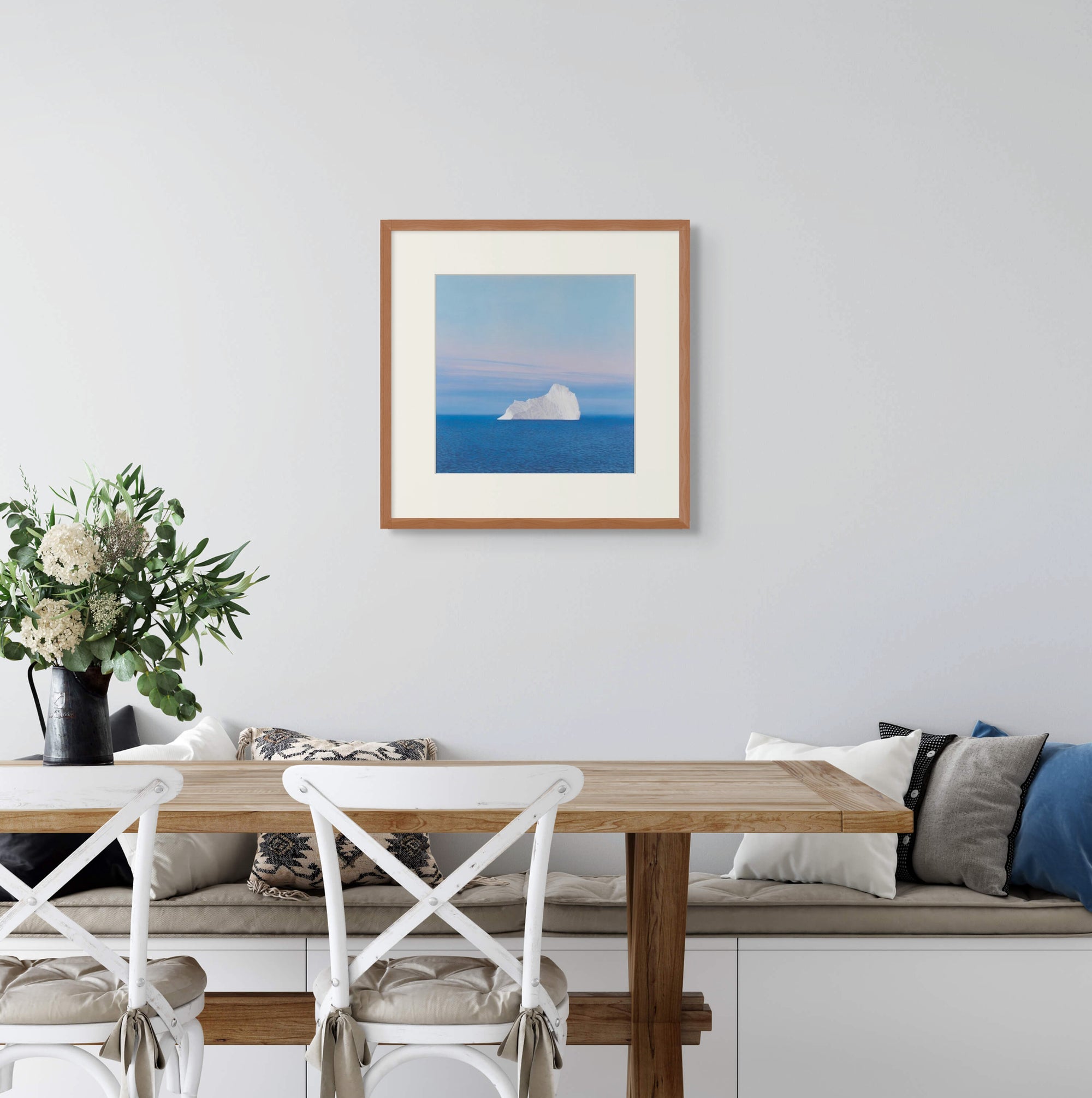 Framed art print of "An Austere Beauty" by Nicholas Jones, shown above a built-in bench in a light-filled kitchen with white chairs and a wooden table set with green and white flowers. Displayed here in size medium.