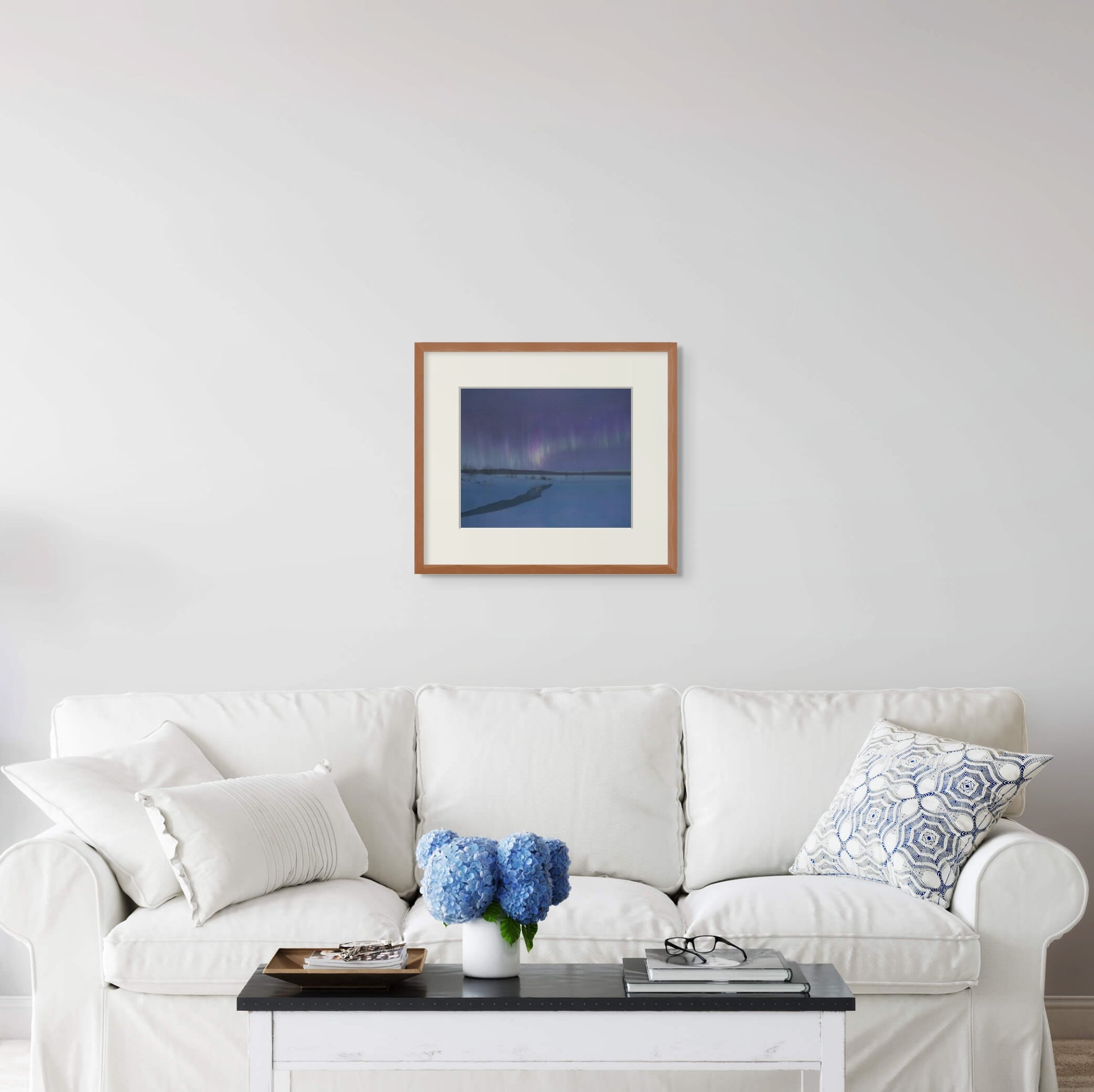 Framed art print of "Aurora Borealis Over Torasieppi" by Nicholas Jones, displayed above a white sofa in a bright, elegant space. A vase of blue hydrangeas sits on the coffee table. Shown here in size medium.
