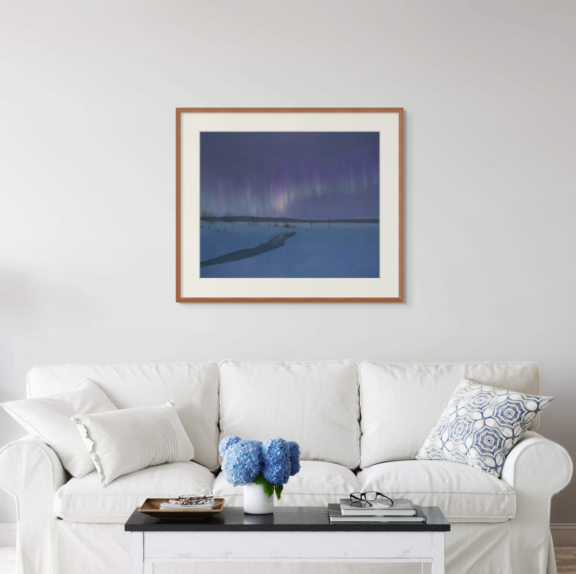 Framed art print of "Aurora Borealis Over Torasieppi" by Nicholas Jones, displayed above a white sofa in a bright, elegant space. A vase of blue hydrangeas sits on the coffee table. Shown here in size extra-large.