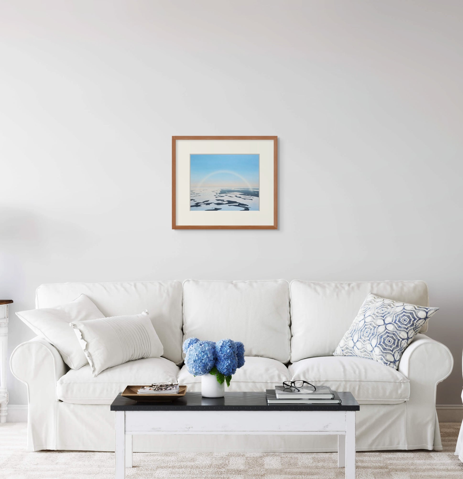 Framed art print of "Fog Bow and Melt Pools" by Nicholas Jones, displayed above a white sofa in a bright, elegant space. A vase of blue hydrangeas sits on the coffee table. Shown here in the size medium.
