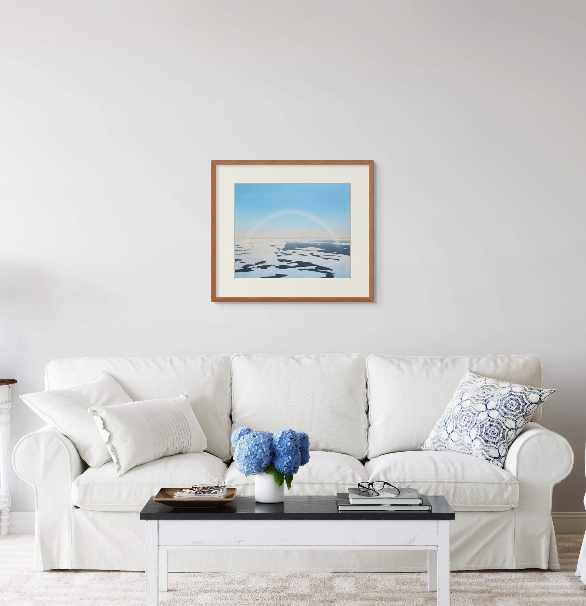 Framed art print of "Fog Bow and Melt Pools" by Nicholas Jones, displayed above a white sofa in a bright, elegant space. A vase of blue hydrangeas sits on the coffee table. Shown here in the size large.