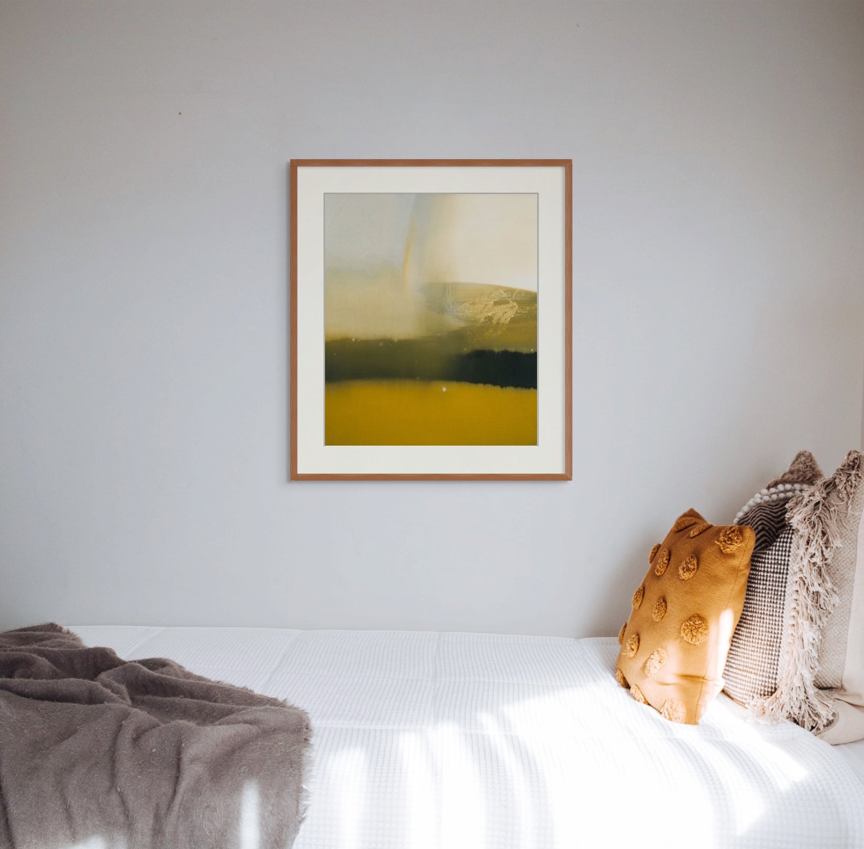 Art print of “Rain's Peculiar Grace” by Nicholas Jones, framed and displayed in a clean white bedroom with golden ochre and pale brown cushions and throw. Sunlight slants in from the window and falls across the white bed cover. Shown here in size extra-large.