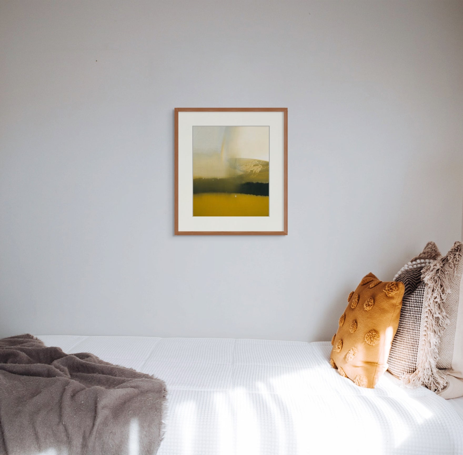 Art print of “Rain's Peculiar Grace” by Nicholas Jones, framed and displayed in a clean white bedroom with golden ochre and pale brown cushions and throw. Sunlight slants in from the window and falls across the white bed cover. Shown here in size large.